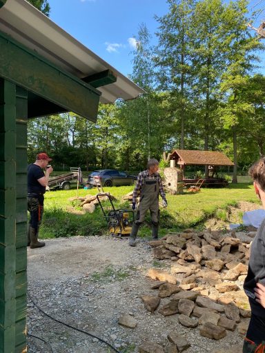Zwei Personen arbeiten an einem Bauprojekt in einem Garten mit Bäumen und einem Spielhaus.