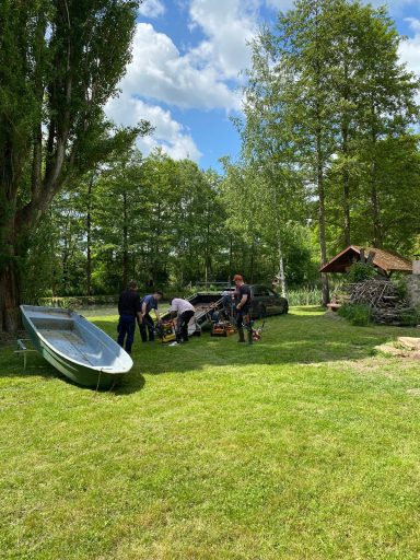 Menschen arbeiten im Freien, umgeben von Bäumen und einem Boot im Gras.