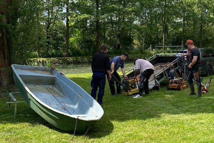 Personen arbeiten im Freien, während ein Boot und Werkzeuge in der Nähe stehen.