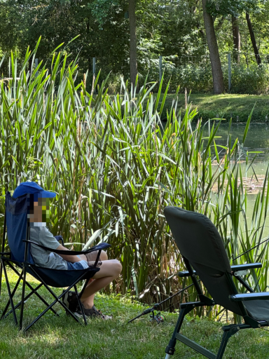 Ein Kind sitzt auf einem Campingstuhl am Ufer eines Teichs, umgeben von Schilfrohr.