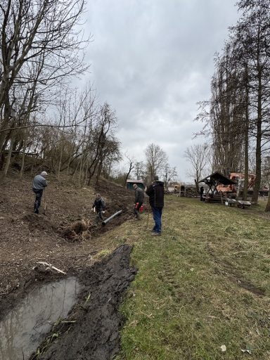 Personen arbeiten im Freien an einem Erdgraben in einer bewaldeten Umgebung.