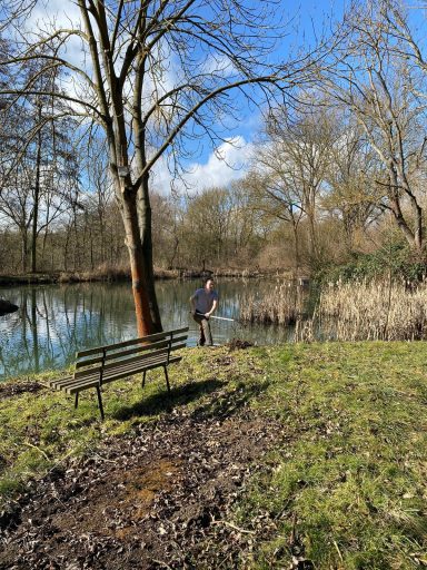 Mann steht am Ufer eines Gewässers, umgeben von Bäumen und einer Bank im Park.