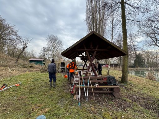 Personen arbeiten unter einem Vordach in einer parkähnlichen Landschaft.