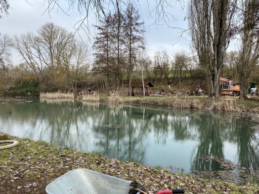 Ruhiger Fluss mit Bäumen und Sträuchern, im Hintergrund sind Menschen zu sehen.