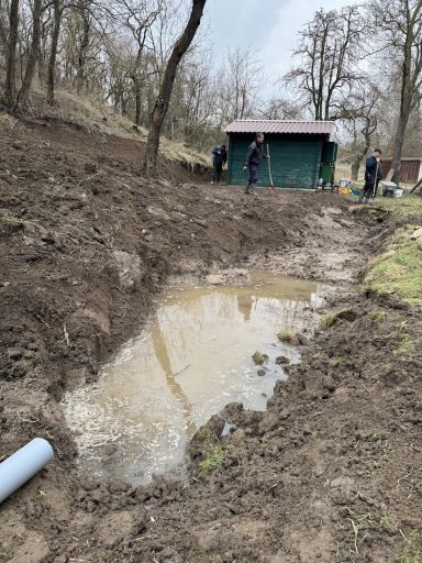 Feuchte Grube im Gelände mit Bauarbeitern und einer Holzhütte im Hintergrund.
