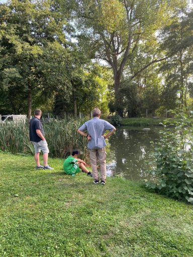 Drei Personen stehen am Ufer eines ruhigen Gewässers, umgeben von grüner Vegetation.
