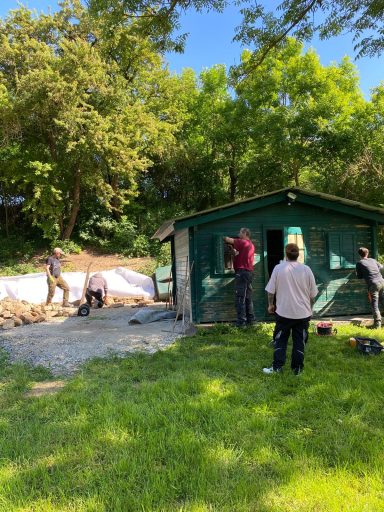 Menschen arbeiten außerhalb an einem grünen Schuppen in einer begrünten Umgebung.