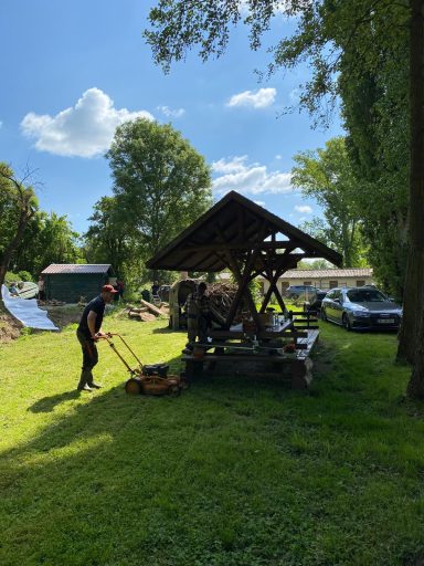 Person schiebt eine Gartenwalze neben einem Holzunterstand im Grünen.