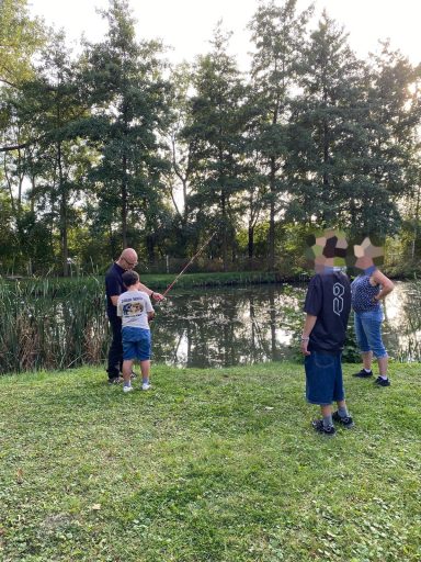 Eine Gruppe von Kindern und Erwachsenen angelt am Ufer eines ruhigen Gewässers.