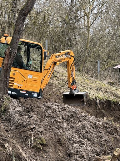Bagger arbeitet an einem Hang, Erde wird bewegt, Bäume im Hintergrund.