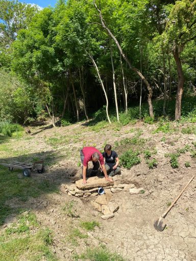 Zwei Personen arbeiten im Garten, umgeben von Bäumen und grüner Vegetation.
