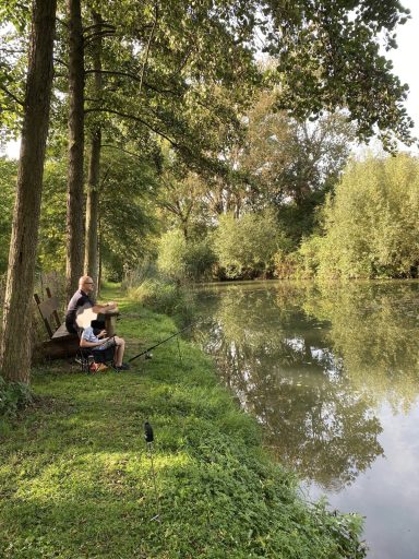 Angler sitzt am Ufer eines ruhigen Gewässers, umgeben von Bäumen und Grün.