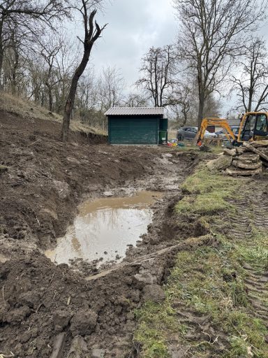 Baugrube mit Wasserpfütze, angrenzender Natur und einem kleinen Gebäude im Hintergrund.