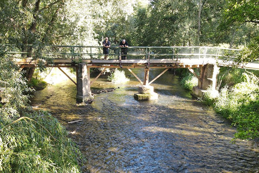 Holzbrücke über einen Bach, zwei Personen stehen darauf, umgeben von Bäumen.