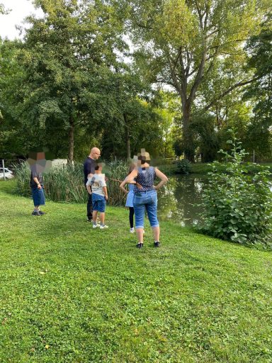 Familie steht am Ufer eines Gewässers, umgeben von Bäumen und Gras.
