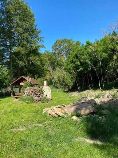 Landschaft mit einer kleinen Hütte, Bäumen und Felsen unter blauem Himmel.