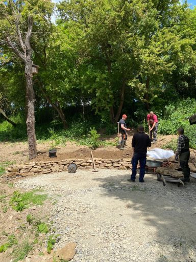 Bauarbeiten in einem grünen Bereich mit Personen, die Materialien bewegen und pflanzen.