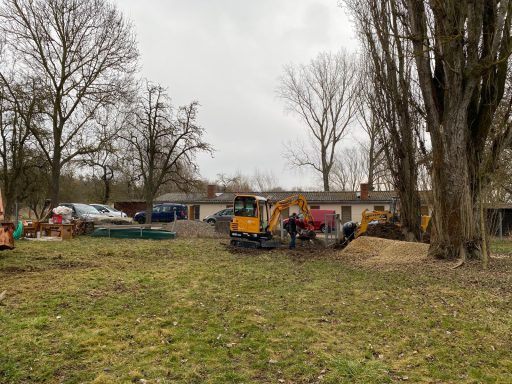 Bauplatz mit Baumaschinen und Fahrzeugen umgeben von Bäumen und Wiese.