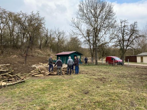 Menschen arbeiten im Freien, Holz wird gesammelt und ein schmales Gebäude steht im Hintergrund.