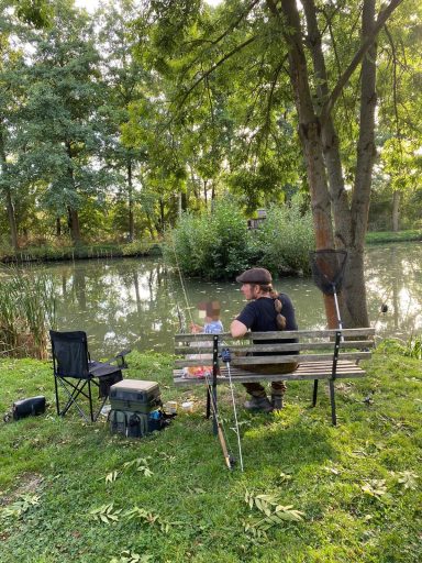 Mann am Ufer eines ruhigen Gewässers, mit Campingstuhl und Tisch, umgeben von Bäumen.