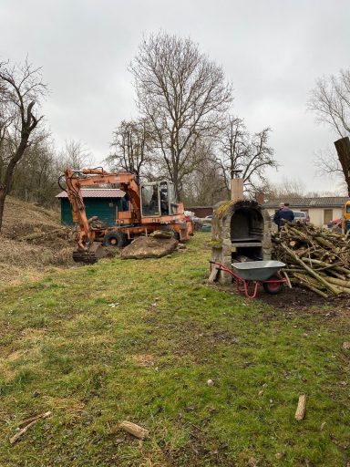 Baugelände mit einem Bagger, einem alten Fahrzeug und Holzstapeln im Hintergrund.