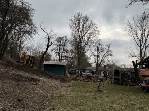 Baulandschaft mit Maschinen, unvollendeten Strukturen und Bäumen unter bewölktem Himmel.
