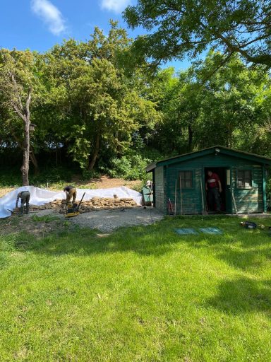 Grünes Gartenhaus neben einem Holzstapel auf einer Wiese mit Bäumen im Hintergrund.